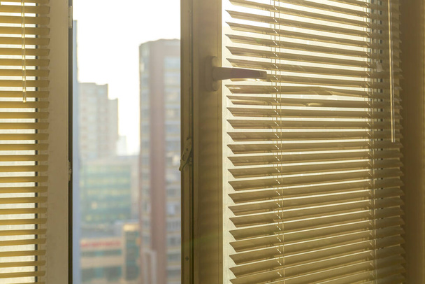 Otevřené okno s okenicemi v bytě s rozmazaným výhledem na město v dálce.Žluté žaluzie a rozmazané domy venku. - Fotografie, Obrázek
