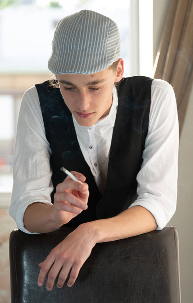 Şapkalı ve yelekli genç bir adam pencerede sigara içiyor. - Fotoğraf, Görsel