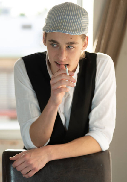 Ein junger Mann in Mütze und Weste raucht am Fenster - Foto, Bild