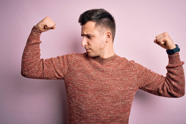 Junger gutaussehender kaukasischer Mann in lässigem Winterpullover über rosa isoliertem Hintergrund, der stolz lächelnde Armmuskeln zeigt. Fitnesskonzept. - Foto, Bild