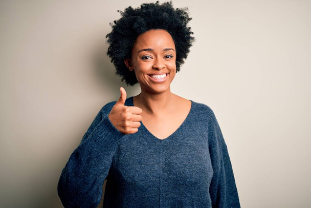 手で幸せな親指アップジェスチャーを行うカジュアルなセーターを着て巻き毛を持つ若い美しいアフリカ系アメリカ人アフロ女性。成功を示すカメラを見て表現を承認する. - 写真・画像