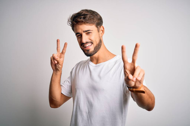 Young handsome man with beard wearing casual t-shirt standing over white background smiling looking to the camera showing fingers doing victory sign. Number two. - Fotó, kép