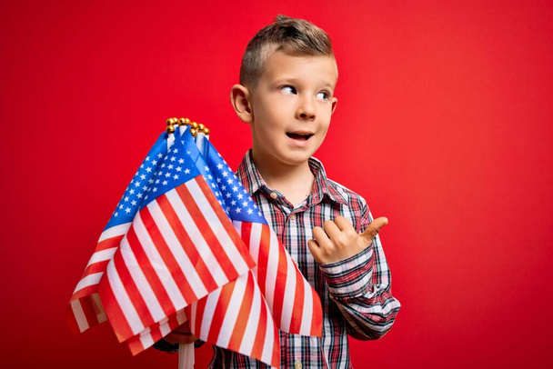 Young little american patriotic caucasian kid holding flag of USA over red background pointing and showing with thumb up to the side with happy face smiling - Fotó, kép