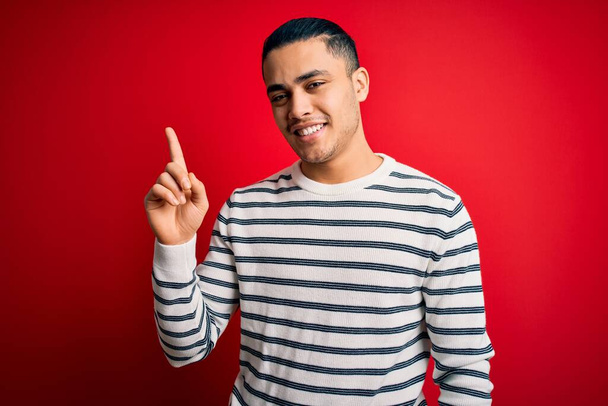 Junger brasilianischer Mann in lässig gestreiftem T-Shirt steht vor isoliertem roten Hintergrund und zeigt mit erhobenem Zeigefinger auf eine erfolgreiche Idee. Aufgeregt und glücklich. Nummer eins. - Foto, Bild