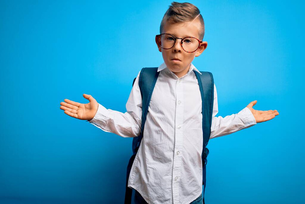 Young little caucasian student kid wearing smart glasses and school bag over blue background clueless and confused expression with arms and hands raised. Doubt concept. - Photo, image