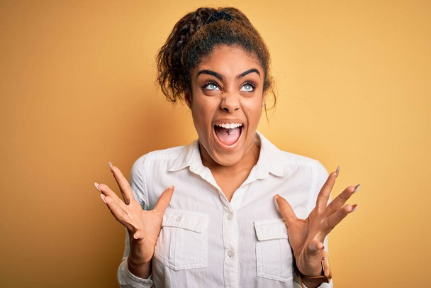 Mladá krásná africká americká dívka v ležérní košili stojící nad žlutým pozadím šílené a šílené křičí a křičí s agresivním výrazem a zvednutými pažemi. Koncept frustrace. - Fotografie, Obrázek