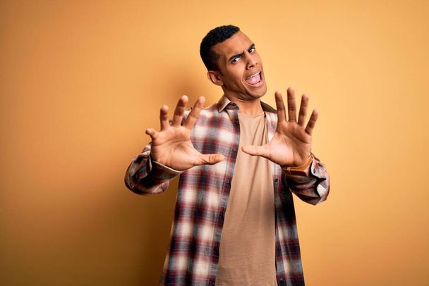 Young handsome african american man wearing casual shirt standing over yellow background afraid and terrified with fear expression stop gesture with hands, shouting in shock. Panic concept. - Photo, Image