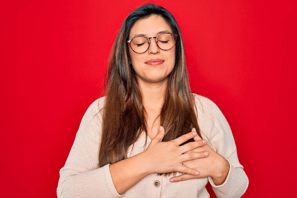 Νεαρή Ισπανίδα έξυπνη γυναίκα με γυαλιά που στέκεται πάνω από κόκκινο απομονωμένο φόντο χαμογελώντας με τα χέρια στο στήθος με κλειστά μάτια και ευγνώμων χειρονομία στο πρόσωπο. Έννοια υγείας. - Φωτογραφία, εικόνα
