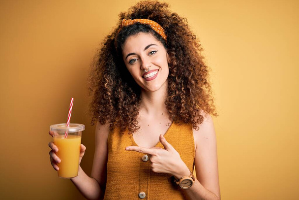 Νεαρή όμορφη γυναίκα με σγουρά μαλλιά και σκουλαρίκια πίνοντας υγιεινό χυμό πορτοκαλιού πολύ χαρούμενος δείχνοντας με το χέρι και το δάχτυλο - Φωτογραφία, εικόνα
