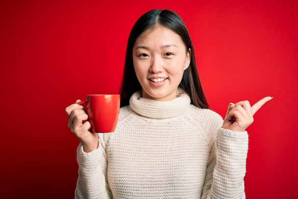 Nuori aasialainen nainen juo kupin kuumaa kahvia yli eristetty punainen tausta erittäin onnellinen osoittaa käsin ja sormella sivulle
 - Valokuva, kuva