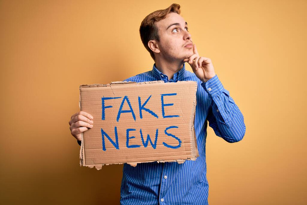 Junge hübsche rothaarige Mann mit Banner mit Fake-News-Nachricht über gelbem Hintergrund ernstes Gesicht denken über Frage mit der Hand am Kinn, nachdenklich über verwirrende Idee - Foto, Bild