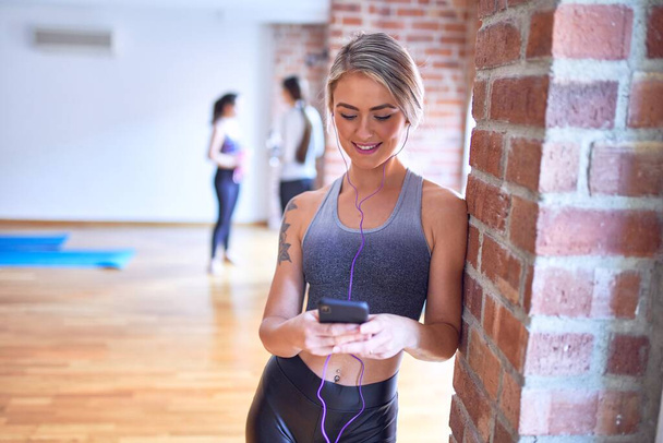 Νεαρή όμορφη αθλήτρια χαμογελά ευτυχισμένη. Στέκεται ακούγοντας μουσική χρησιμοποιώντας ακουστικά και smartphone μετά το μάθημα γιόγκα στο γυμναστήριο - Φωτογραφία, εικόνα