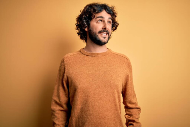 Young handsome man with beard wearing casual sweater standing over yellow background smiling looking to the side and staring away thinking. - Photo, Image
