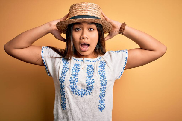 Joven hermosa chica asiática vistiendo casual camiseta y sombrero de pie sobre fondo amarillo Loco y asustado con las manos en la cabeza, miedo y sorprendido de choque con la boca abierta
 - Foto, imagen