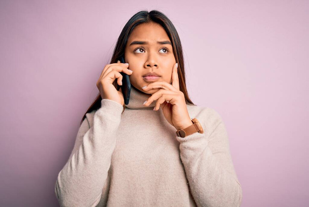 Mladá asijská dívka vede rozhovor na smartphonu přes izolované růžové pozadí vážný obličej přemýšlet o otázce, velmi zmatený nápad - Fotografie, Obrázek