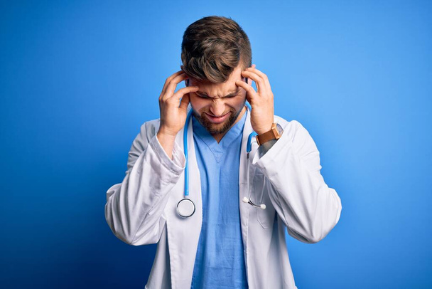 Young blond doctor man with beard and blue eyes wearing white coat and stethoscope with hand on head for pain in head because stress. Suffering migraine. - Foto, immagini