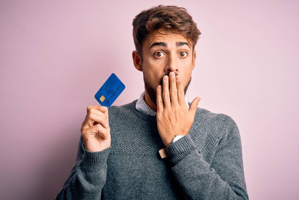 Mladý zákazník muž s vousy držící kreditní kartu pro platbu přes růžové pozadí kryt úst s rukou šokován hanbou za omyl, vyjádření strachu, strach v tichu, tajný koncept - Fotografie, Obrázek