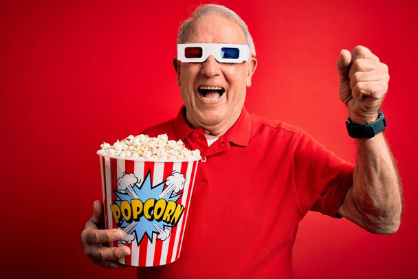 Siwy starszy mężczyzna w okularach 3D i jedzący popcorn na czerwonym tle krzycząc dumny i świętując zwycięstwo i sukces bardzo podekscytowany, dopingujące emocje - Zdjęcie, obraz