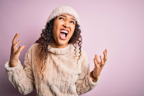 Beautiful woman with curly hair wearing casual sweater and wool cap over pink background crazy and mad shouting and yelling with aggressive expression and arms raised. Frustration concept. - Fotó, kép