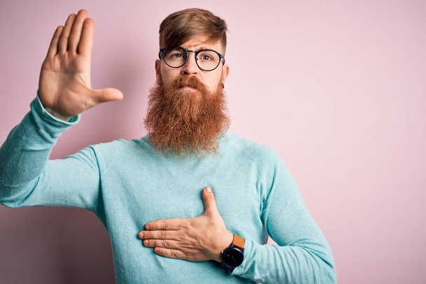 Bello irlandese rossa uomo con barba indossare occhiali su sfondo rosa isolato giurando con mano sul petto e palmo aperto, facendo un giuramento di fedeltà
 - Foto, immagini