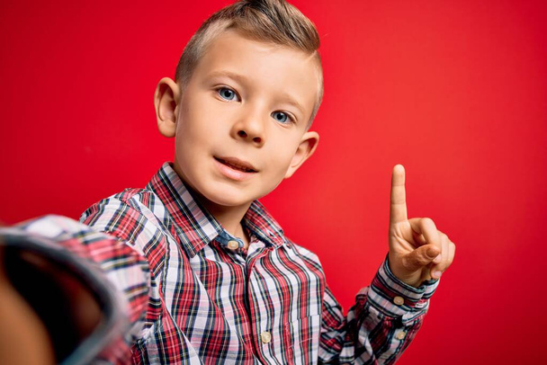 lähikuva nuoresta valkoihoisesta sinisilmäisestä pojasta ottamassa selfiekuvaa punaisella taustalla yllättynyt ideasta tai kysymyksestä, joka osoittaa sormella onnellisia kasvoja, numero yksi
 - Valokuva, kuva