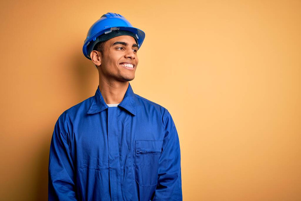 Νεαρός όμορφος Αφροαμερικάνος εργάτης με μπλε στολή και κράνος να κοιτάει από την άλλη πλευρά με χαμόγελο στο πρόσωπο, φυσική έκφραση. Γελάς σίγουρος. - Φωτογραφία, εικόνα