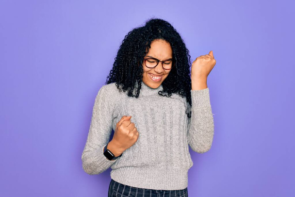 Jeune femme afro-américaine portant un pull décontracté et des lunettes sur fond violet célébrant surpris et étonnés pour le succès avec les bras levés et les yeux fermés. Concept gagnant
. - Photo, image