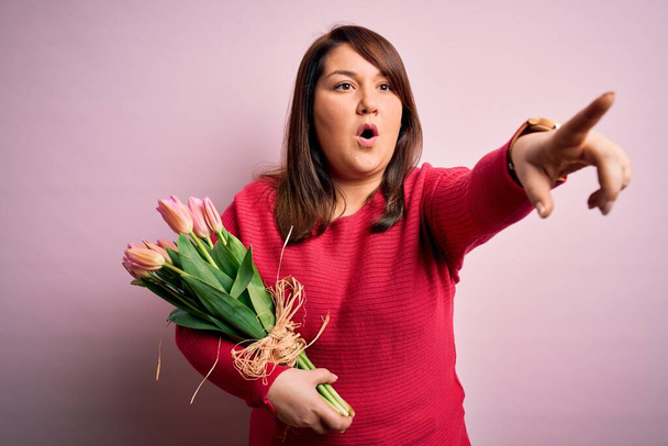 Krásná plus velikost žena drží romantickou kytici přírodních tulipánů květiny přes růžové pozadí ukazuje s prstem překvapený dopředu, otevřená ústa užaslý výraz, něco na přední - Fotografie, Obrázek