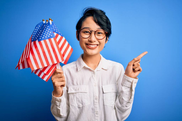 Молода красива азіатська патріотична дівчина тримає єдині державні прапори, святкуючи День незалежності, здивована ідеєю або питанням, що вказує пальцем на щасливе обличчя, номер один
 - Фото, зображення