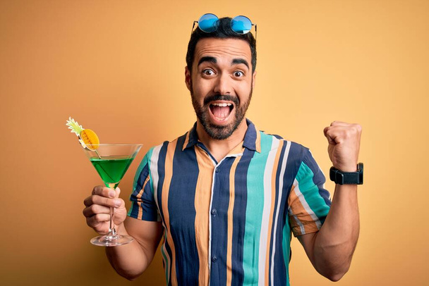 Junger gutaussehender Touristenmann mit Bart im Urlaub trägt Sommerhemd, trinkt Cocktail und schreit stolz und feiert Sieg und Erfolg sehr aufgeregt, jubelnde Emotionen - Foto, Bild