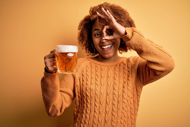 Jonge mooie Afro-Amerikaanse afro vrouw met krullend haar drinken pot bier met gelukkig gezicht glimlachen doen ok teken met de hand op oog kijken door vingers - Foto, afbeelding