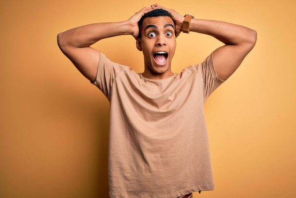 Young handsome african american man wearing casual t-shirt standing over yellow background Crazy and scared with hands on head, afraid and surprised of shock with open mouth - Foto, Imagem