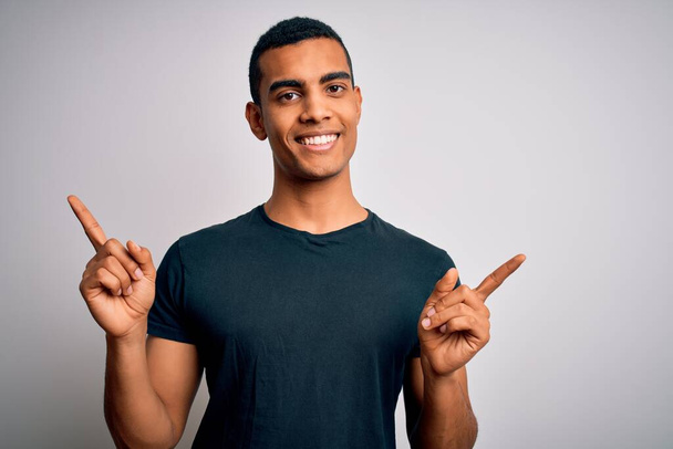 Nuori komea afrikkalainen amerikkalainen mies päällään rento t-paita seisoo valkoisella taustalla hymyillen luottavainen sormin eri suuntiin. Kopioi tilaa mainokselle
 - Valokuva, kuva