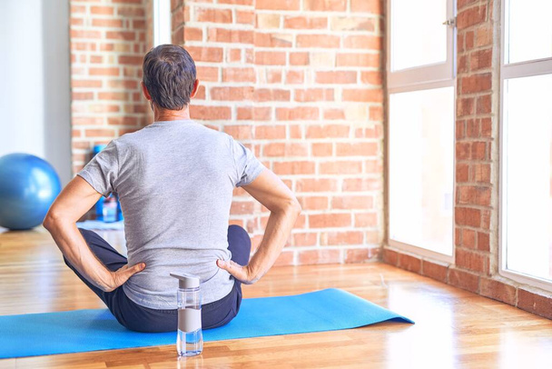 Esportista bonito de meia idade sentado no tapete fazendo alongamento exercício de ioga no ginásio de pé para trás olhando para longe com os braços no corpo
 - Foto, Imagem