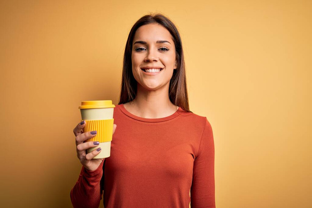Jeune belle femme brune buvant tasse de café à emporter sur fond jaune avec un visage heureux debout et souriant avec un sourire confiant montrant les dents
 - Photo, image