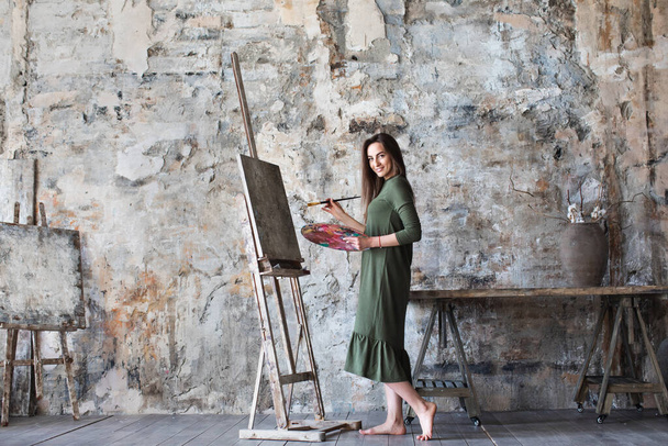 Photo of a woman dressed in a green dress painting in an art studio and smiling. Hobby. - Foto, Imagen