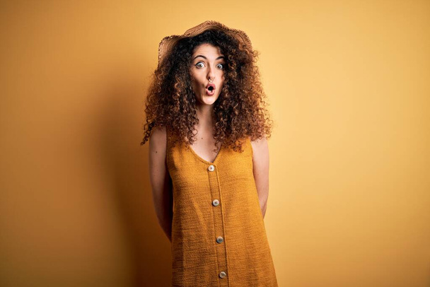 Beautiful brunette woman on vacation with curly hair and piercing wearing hat and dress afraid and shocked with surprise expression, fear and excited face. - Photo, Image
