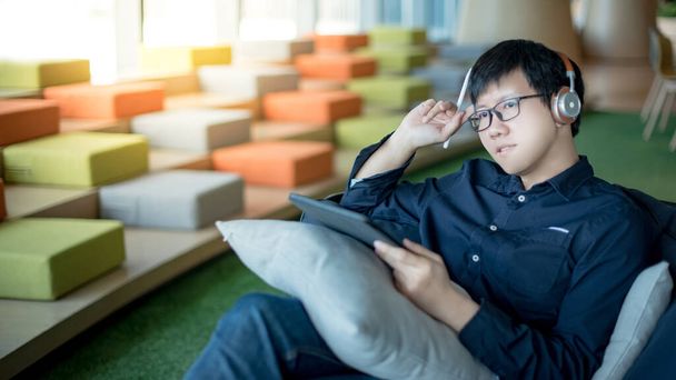 ヘッドフォンを持つアジアのビジネスマンは、自宅のオフィスで快適なソファでデジタルタブレットで音楽を聴いています。メガネをかけた男がモバイルアプリでオンライン会議をしている。家のコンセプトからの仕事 - 写真・画像