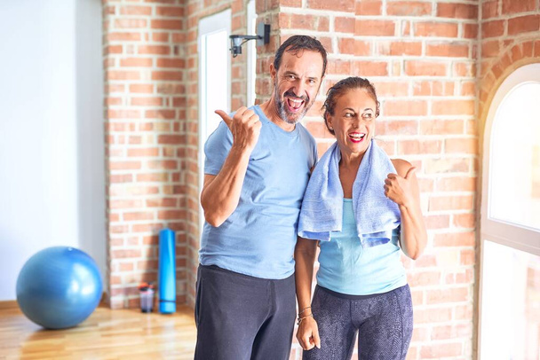 Middelbare leeftijd sportief paar staan voor doen oefening in de sportschool glimlachen met een vrolijk gezicht kijken en wijzen naar de zijkant met duim omhoog. - Foto, afbeelding