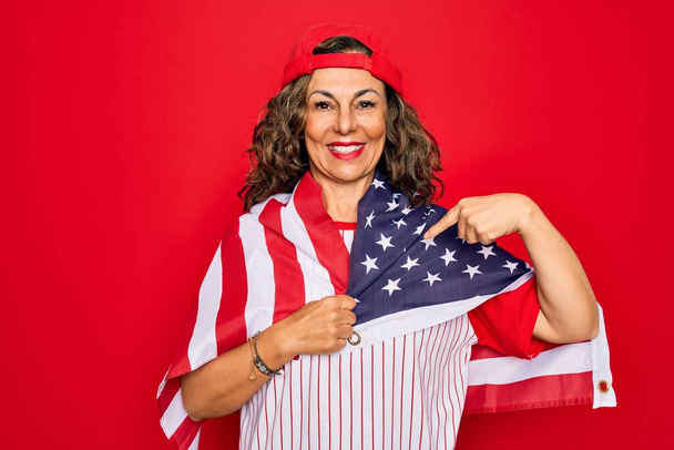 Patriotische Frau mittleren Alters trägt Baseballausrüstung mit US-Flagge und zeigt überrascht mit dem Finger auf sich selbst - Foto, Bild
