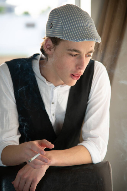 Ένας νεαρός με καπέλο και γιλέκο καπνίζει δίπλα στο παράθυρο. - Φωτογραφία, εικόνα