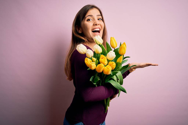 Молодая блондинка держит в руках романтический букет из желтых тюльпанов цветы на розовом фоне, указывая в сторону с открытыми руками ладони показывая копировальное пространство, представляя рекламу улыбаясь взволнованный счастливым
 - Фото, изображение