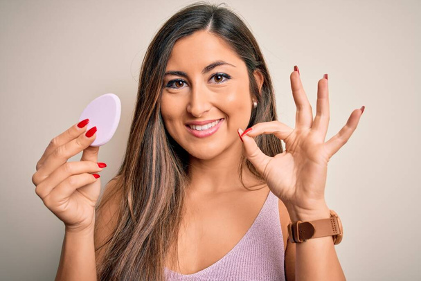 Young beautiful woman using beauty sponge with facial makeup over pink background doing ok sign with fingers, excellent symbol - Foto, Imagem