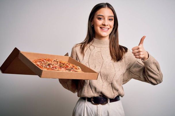 Young beautiful brunette woman holding box with italian pizza over white background happy with big smile doing ok sign, thumb up with fingers, excellent sign - Valokuva, kuva