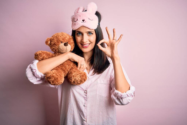 Junge brünette Frau mit blauen Augen trägt Pyjama umarmt Teddybär Stofftier tut ok Zeichen mit den Fingern, hervorragendes Symbol - Foto, Bild