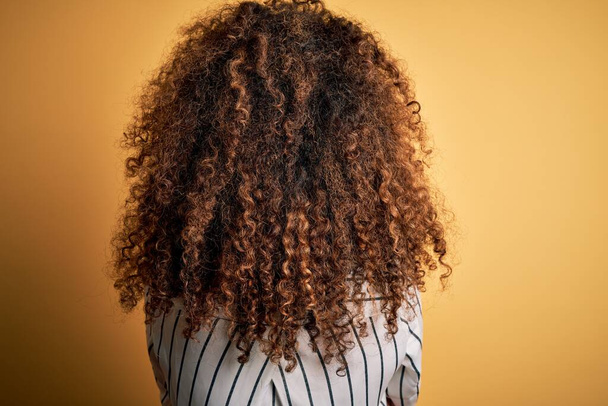 Young beautiful woman with curly hair and piercing wearing striped shirt and glasses standing backwards looking away with crossed arms - Фото, изображение