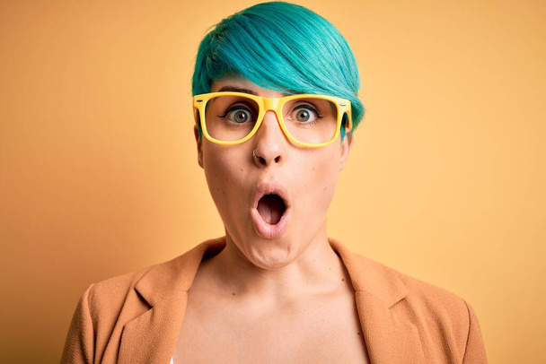 Jovem mulher bonita com cabelo azul moda vestindo óculos casuais sobre fundo amarelo assustado em choque com um rosto surpresa, com medo e animado com expressão de medo
 - Foto, Imagem