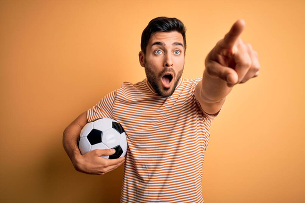 Όμορφος παίκτης με γενειάδα παίζει ποδόσφαιρο κρατώντας μπάλα ποδοσφαίρου πάνω από κίτρινο φόντο Δείχνοντας με το δάχτυλο έκπληκτος μπροστά, ανοιχτό στόμα κατάπληκτος έκφραση, κάτι στο μπροστινό μέρος - Φωτογραφία, εικόνα