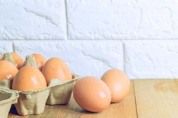 Куриные яйца в бумажном лотке и яйца, помещенные на деревянный стол на кухне с кирпичным фоном - Фото, изображение