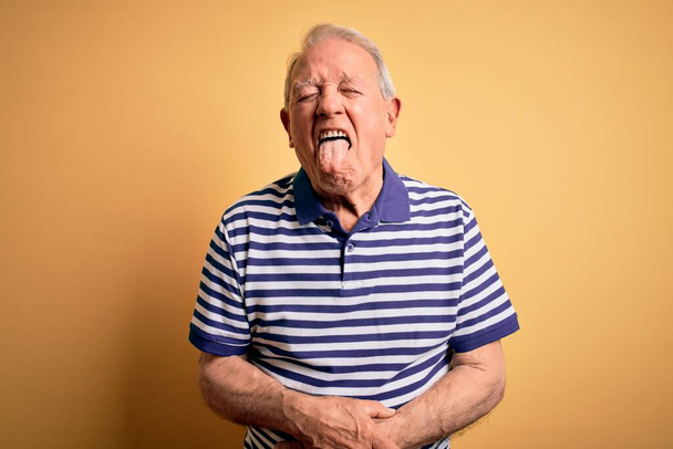 Grey haired senior man wearing casual navy striped t-shirt standing over yellow background sticking tongue out happy with funny expression. Emotion concept. - Photo, Image
